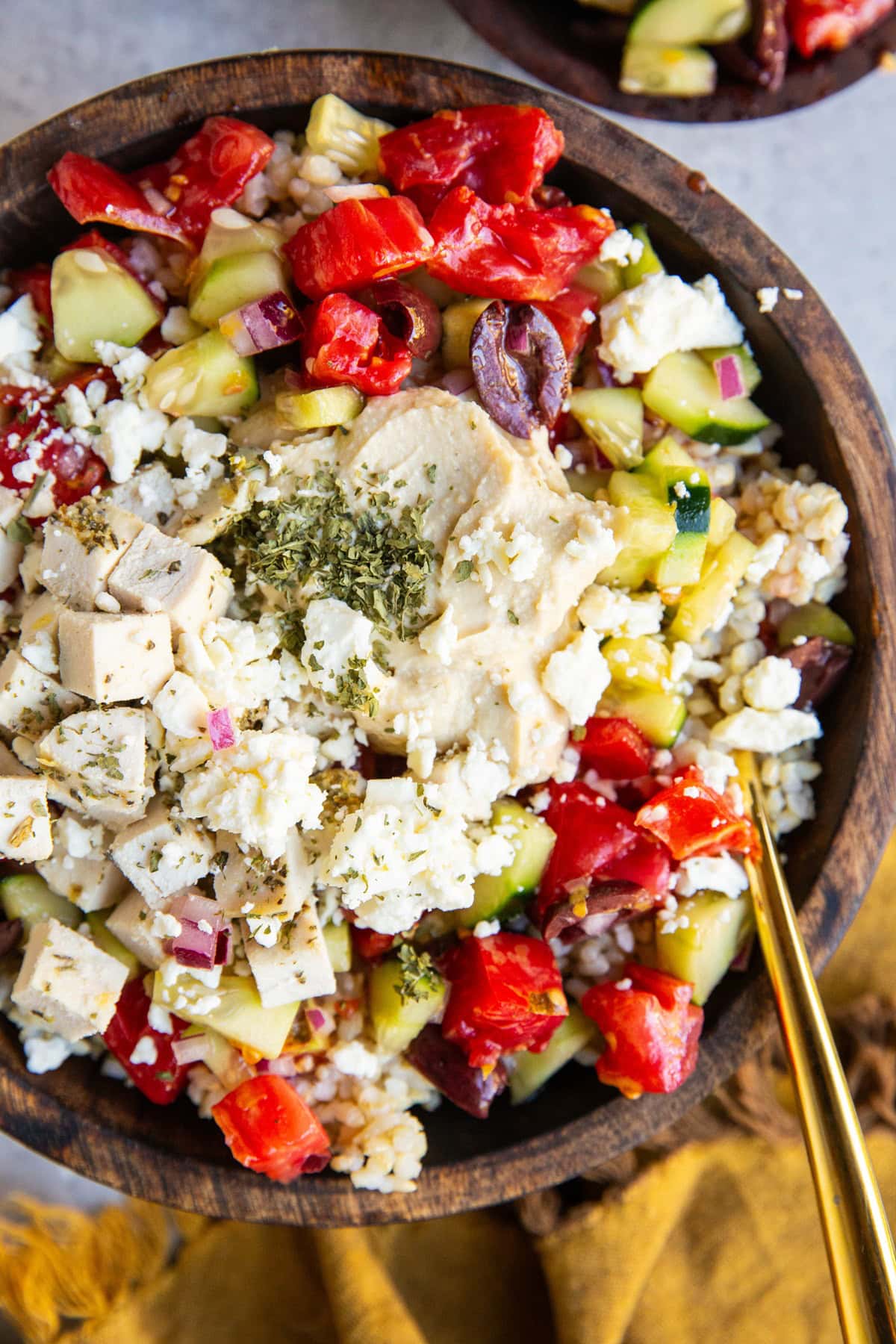 Big wooden bowl of Mediterranean chicken, hummus, and cucumber tomato salad with a golden napkin and spoon.