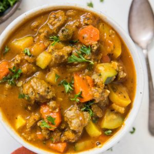 Big bowl of soup with fresh vegetables and meat. A red napkin, spoon, and fresh herbs to the side of the bowl.