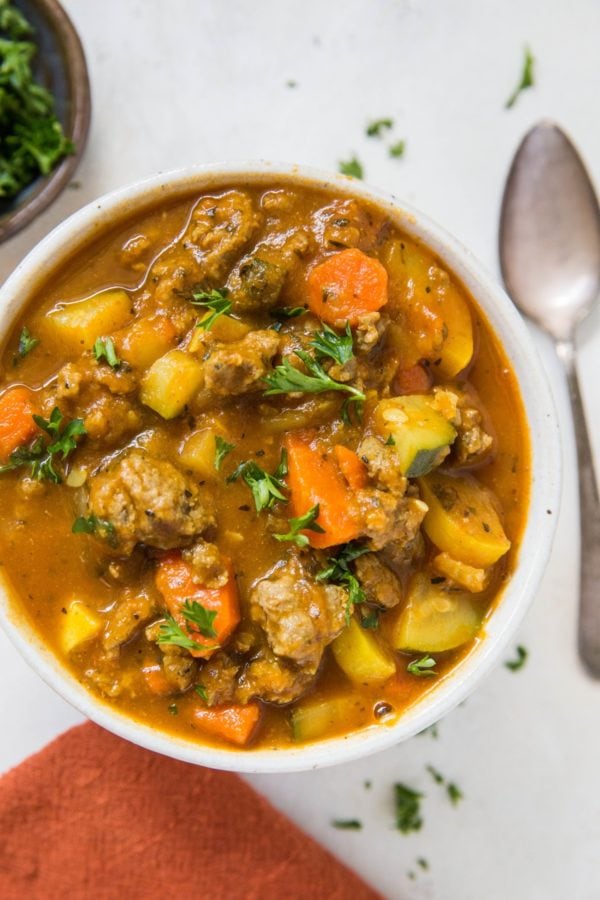 Big bowl of soup with fresh vegetables and meat. A red napkin, spoon, and fresh herbs to the side of the bowl.