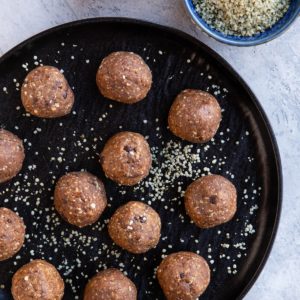 Black plate full of energy balls sprinkled with hemp seeds. A bowl and measuring cup of oats and hemp seeds to the side.