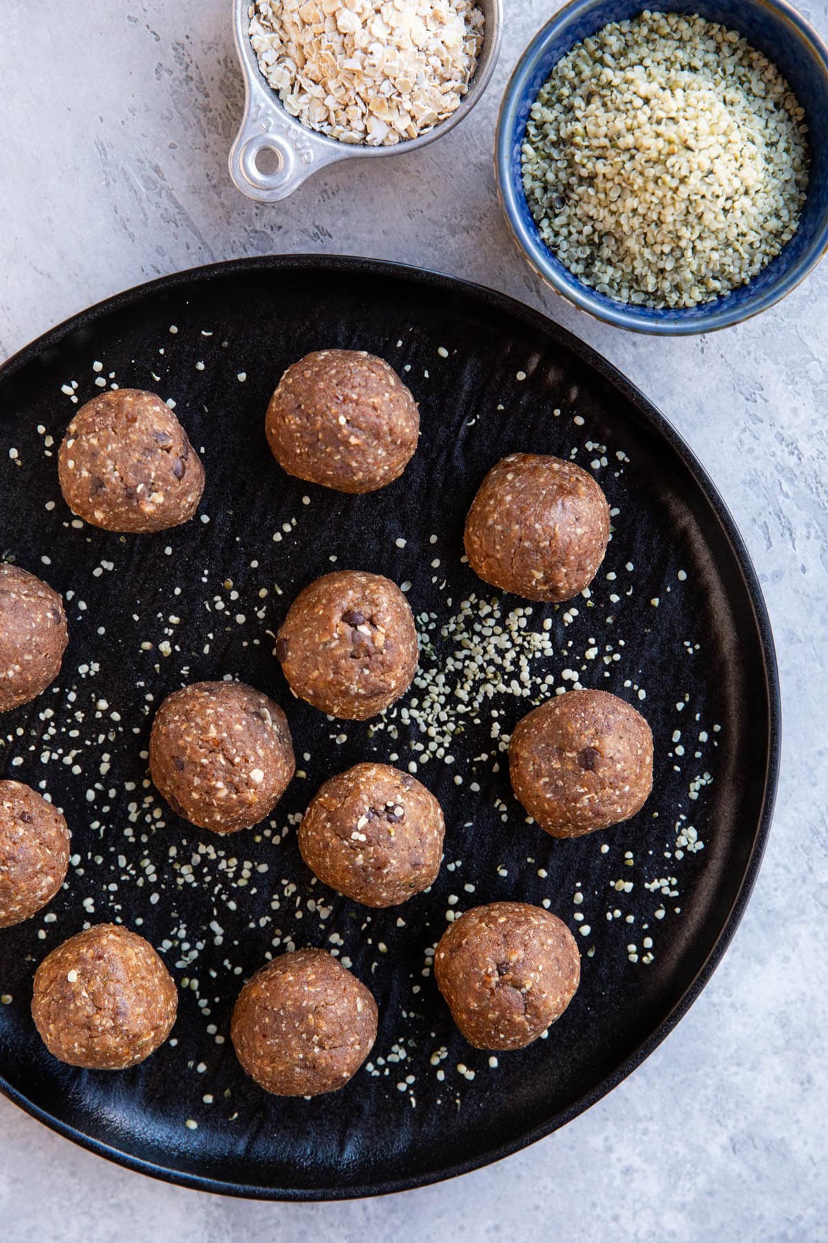 Black plate full of energy balls sprinkled with hemp seeds. A bowl and measuring cup of oats and hemp seeds to the side.