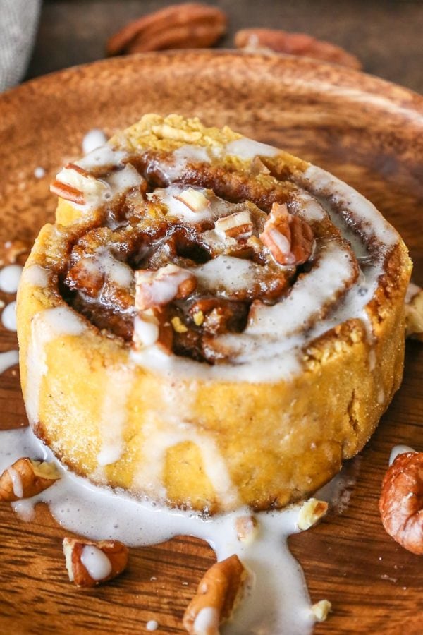 Large pumpkin cinnamon roll with pecans and glaze on a wooden plate, ready to eat.