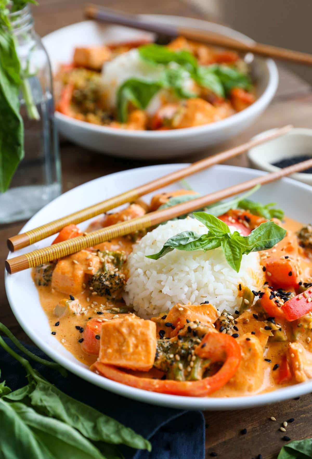 Two bowls of salmon curry with scoops of rice in the center and fresh basil all around. Chop sticks and a blue napkin for serving.