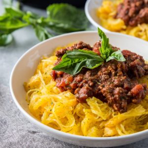 Big white bowl of spaghetti squash with red meat sauce on top and a basil leaf.