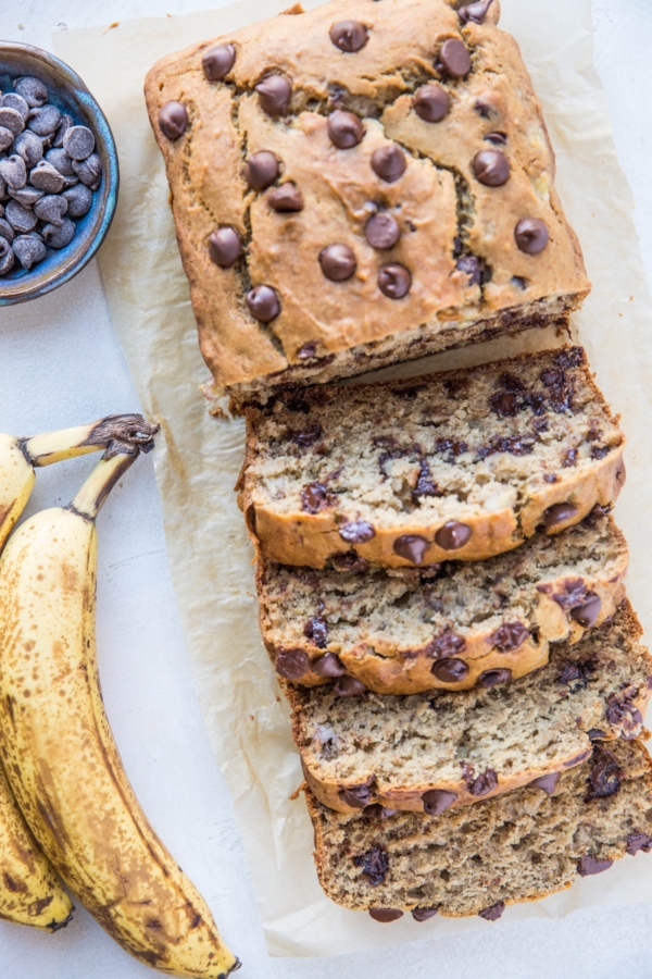 Loaf of banana bread cut into slices on a sheet of parchment paper with ripe bananas to the side.