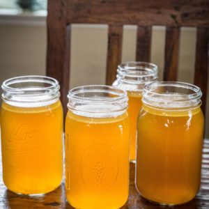 Four large jars full of bone broth cooling on a wooden table with a wood chair in the background.