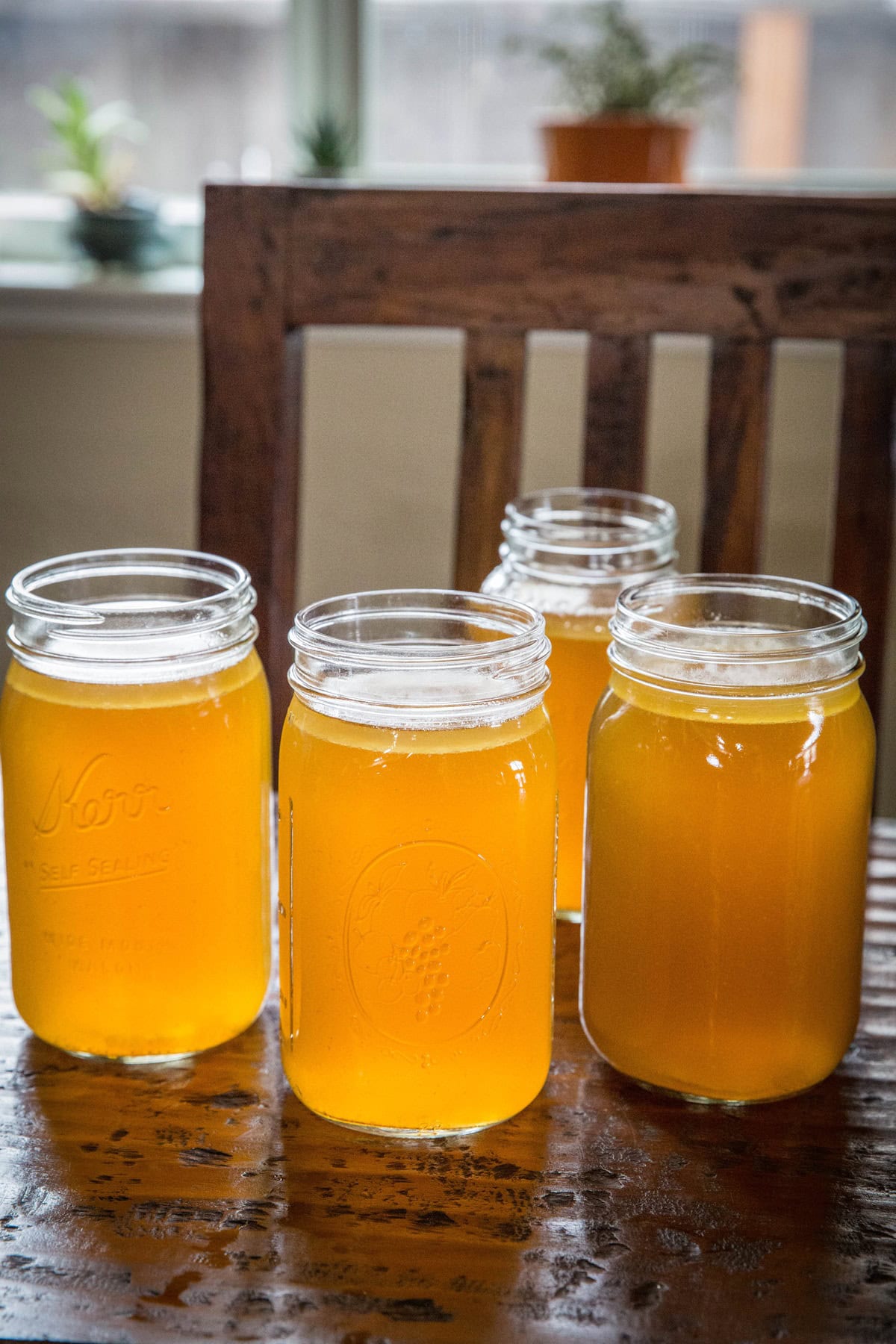 Four large jars full of bone broth cooling on a wooden table with a wood chair in the background.