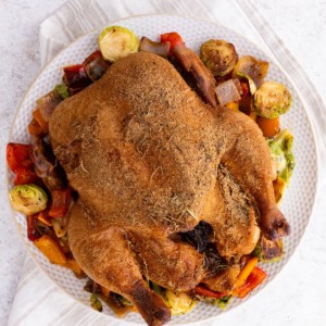 Whole smoked chicken on a plate on top of roasted vegetables, fresh off the smoker grill and ready to serve.