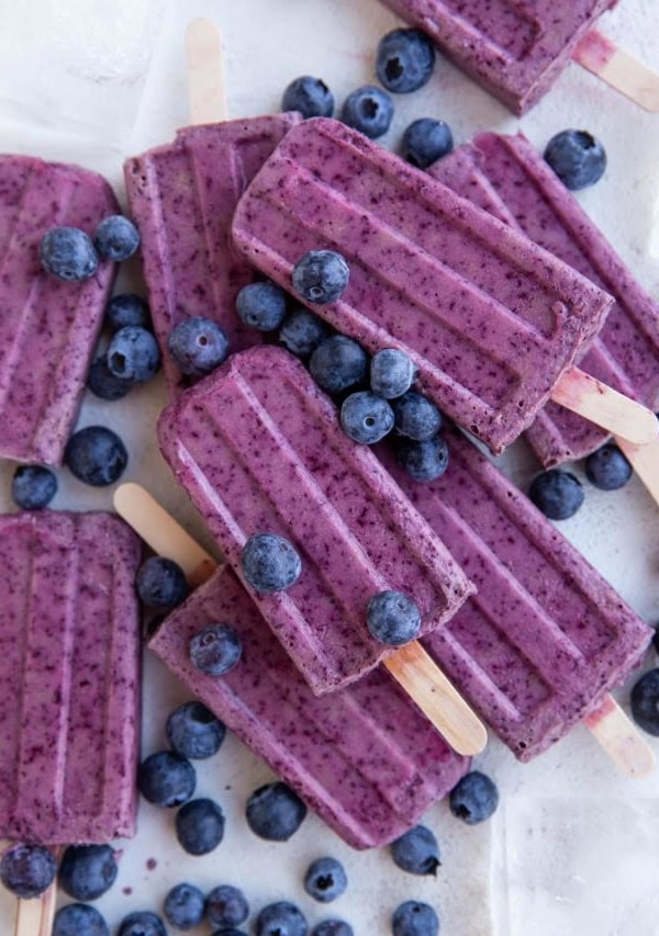 Pile of blueberry coconut milk popsicles on a white background with ice and fresh blueberries all around.