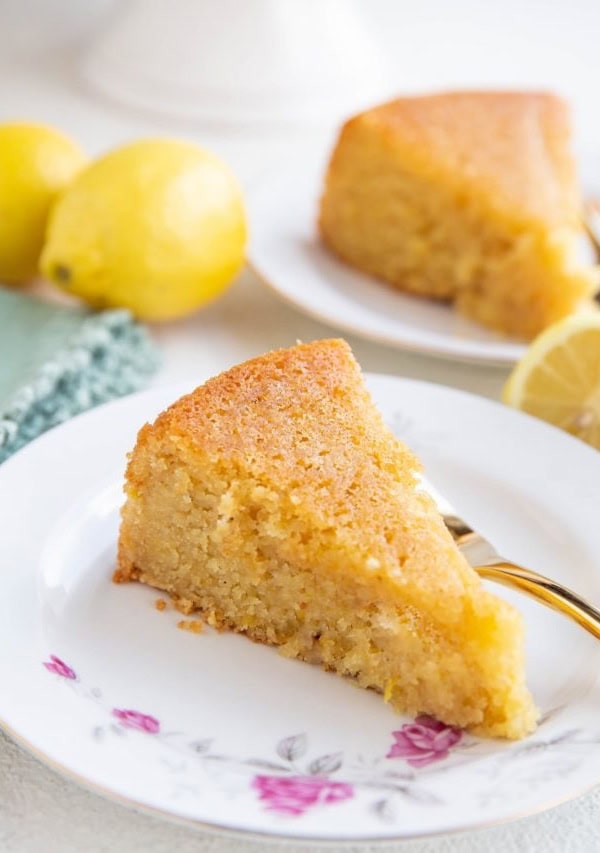 Two slices of lemon cake on two plates with fresh lemons all around.