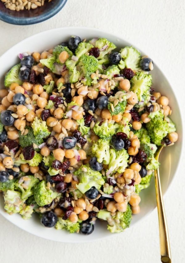 Big white bowl of broccoli chickpea salad with a gold spoon and a bowl of sunflower seeds.