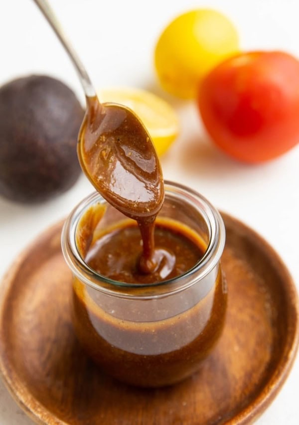 Spoon drizzling balsamic vinaigrette in a jar with fresh produce in the background.