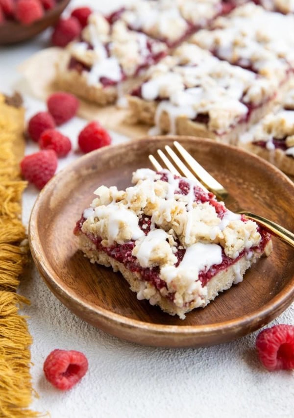 Wooden plate with a slice of raspberry crumb bar and a gold fork. More crumb bars in the background.