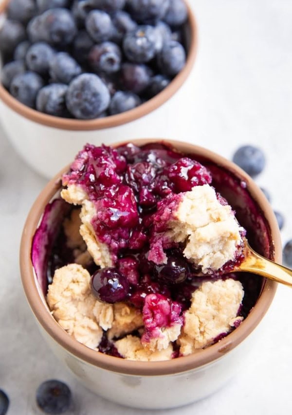 White ramekin with blueberry cobbler and a bowl of fresh blueberries in the background.