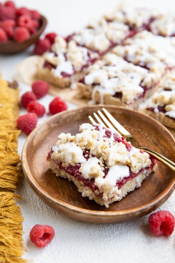 Wooden plate with a slice of raspberry crumb bar and a gold fork. More crumb bars in the background.