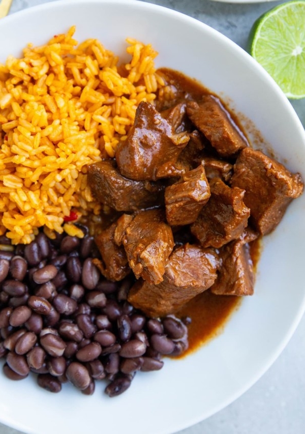 White bowl of Chile Colorado, rice, beans, and a fresh lime to the side.