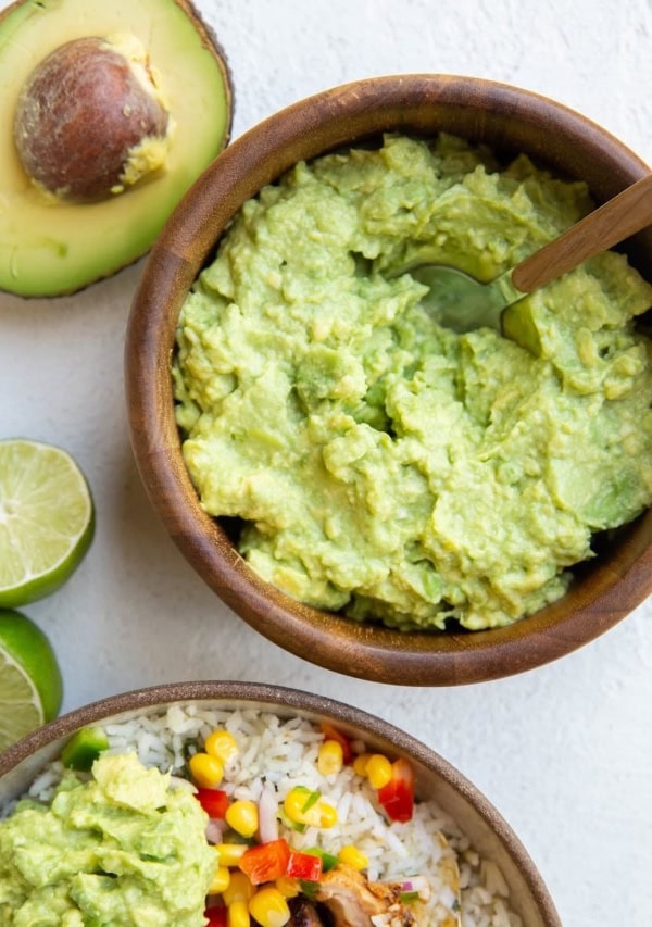 Wooden bowl of guacamole with a chicken burrito bowl to the side with guacamole on top.