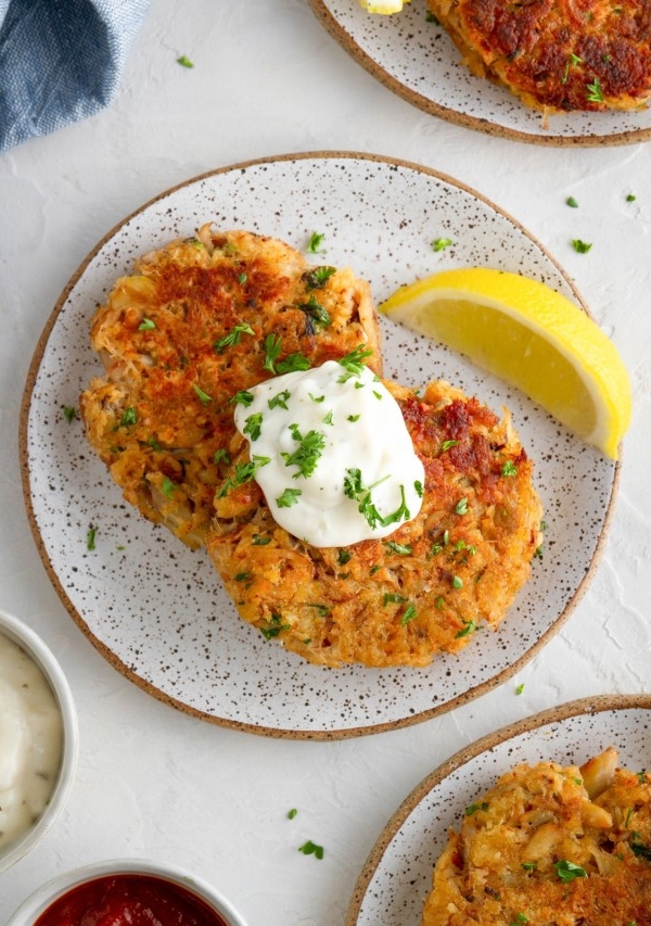 Three small appetizer plates with crab cakes on top and tartar sauce and cocktail sauce to the side.