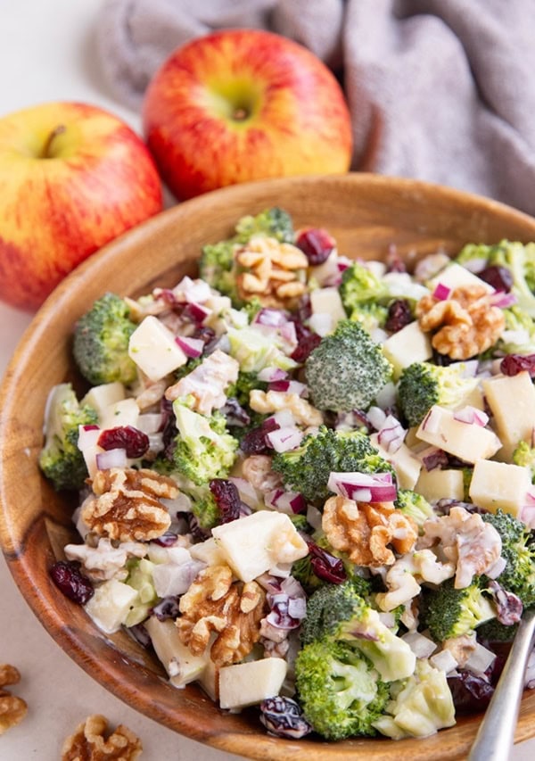 Apple broccoli salad in w wooden bowl with a silver serving spoon and fresh apples in the background.