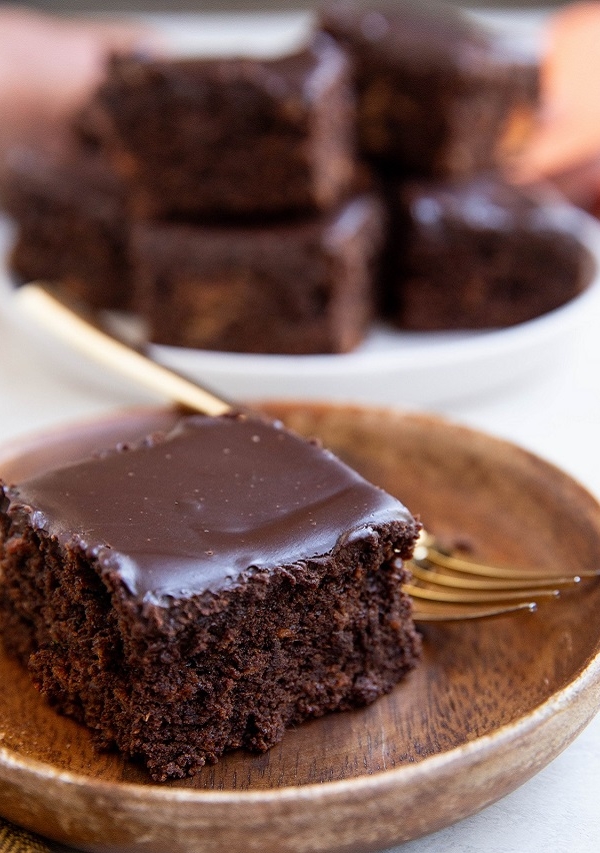 Slice of sweet potato chocolate cake on a wooden plate with a white plate of slices of cake in the background.