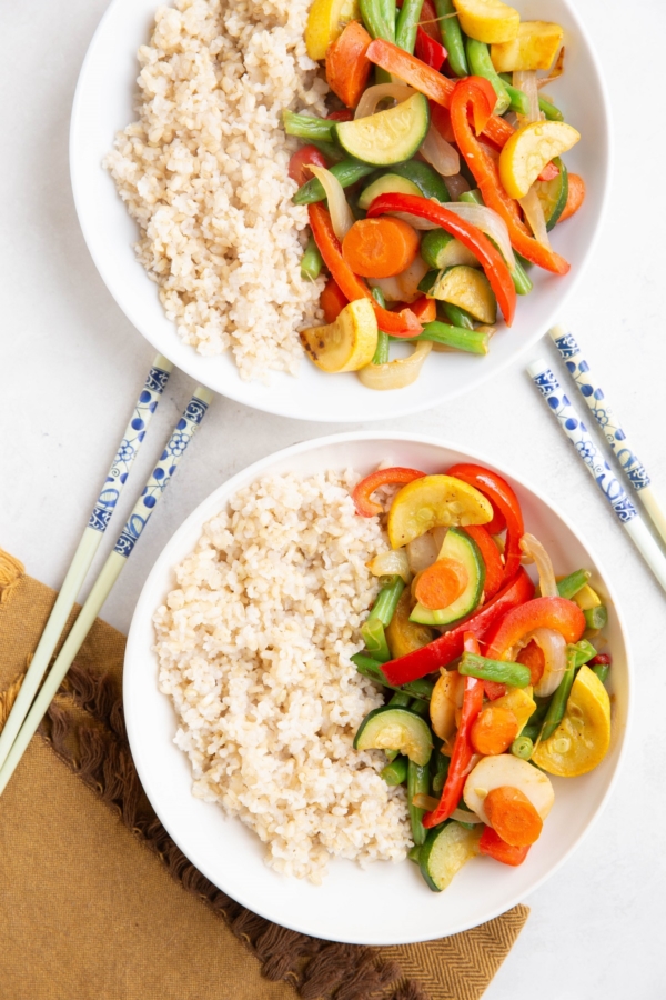 Two white bowls of stir-fry vegetables with brown rice and chopsticks.
