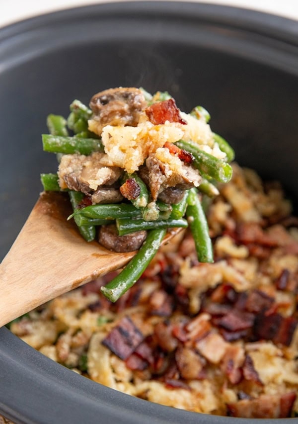 Wooden spoon scooping green bean casserole out of a crock pot.