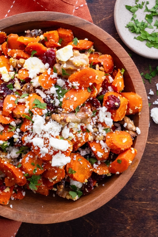 Maple Roasted Carrots in a wooden bowl with feta, dried cranberries and walnuts and a small bowl of parsley to the side.