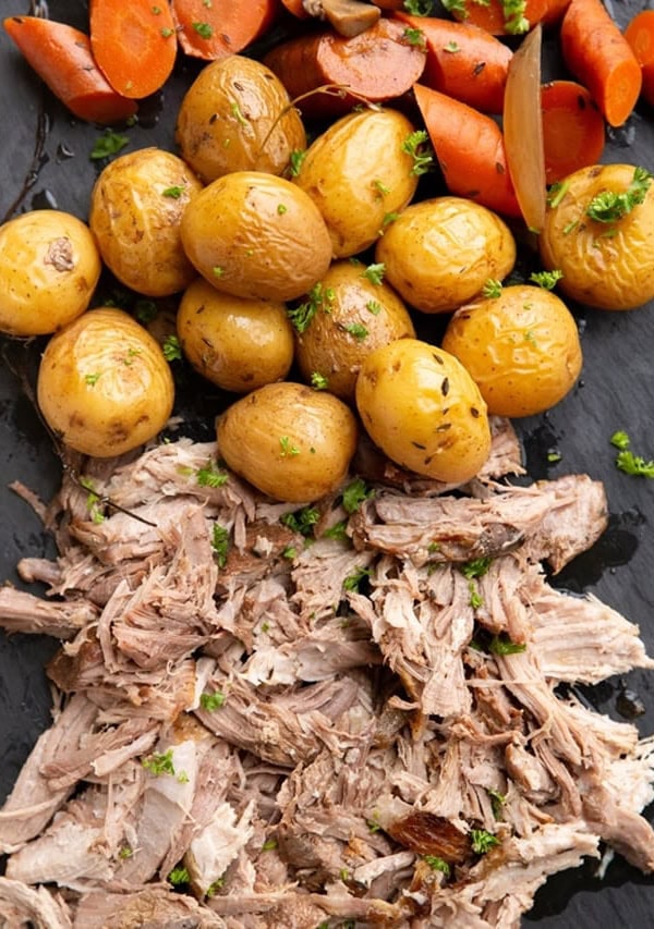 Black serving platter with shredded pork roast, yellow potatoes and carrots.