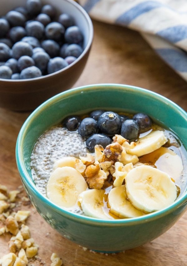 Teal bowl of chia pudding with sliced bananas, walnuts, and fresh berries on top. Chopped walnuts to the side, a bowl of fresh blueberries in the background, and a blue striped napkin.