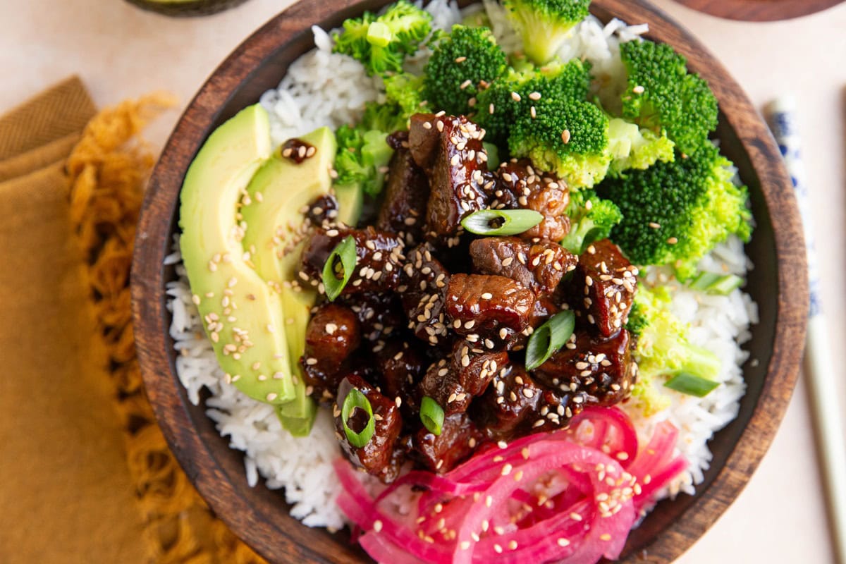 Wooden bowl of steak bites with rice and broccoli and avocado with chopsticks and a napkin to the side.