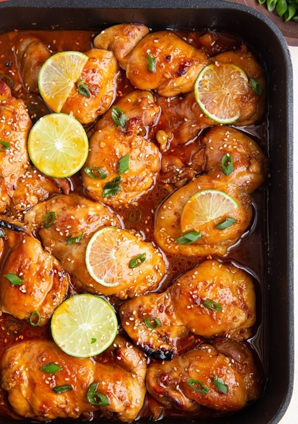 Casserole dish full of Korean BBQ chicken, sprinkled with green onions and lime slices.