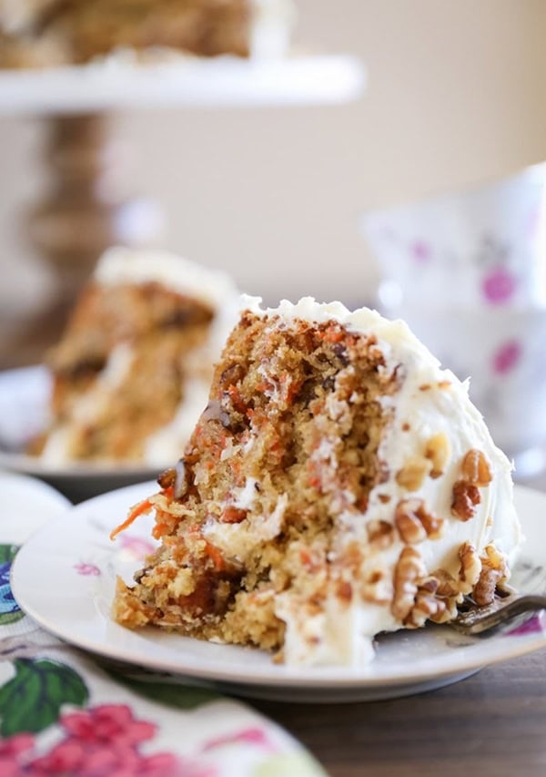 Slice of carrot cake on a plate with carrot cake in the background.