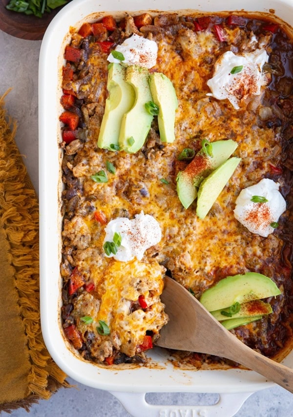 White casserole dish full of cheesy ground turkey casserole with sliced avocado and sour cream on top.