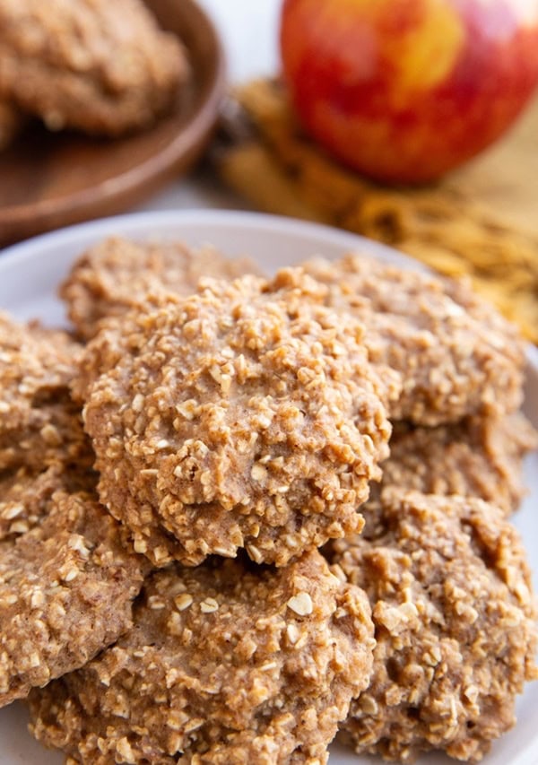 White plate with oatmeal cookies on top and a wooden plate in the background with more cookies and a fresh apple.