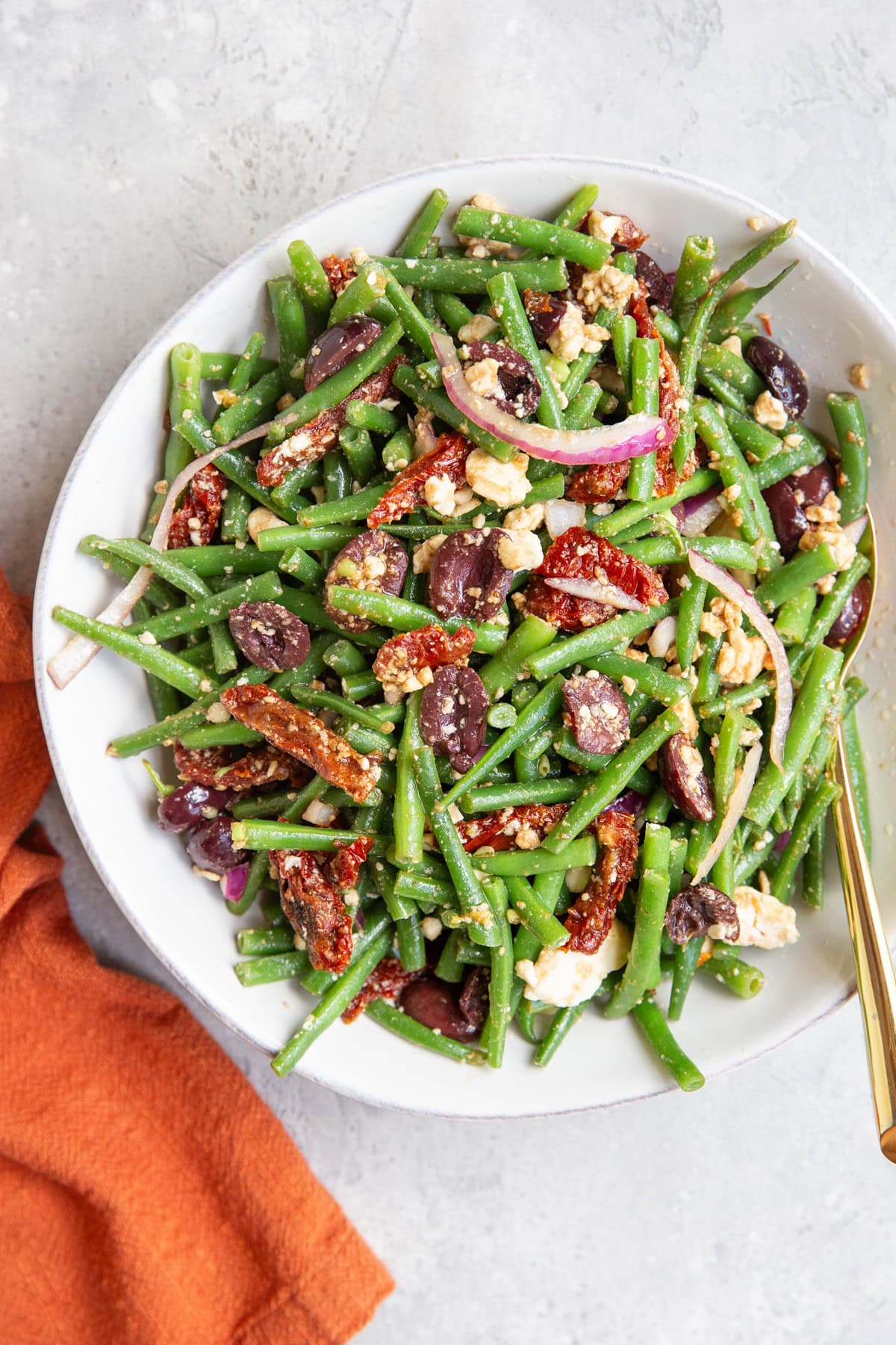 Green bean salad in a white bowl with a golden spoon and a red napkin to the side.