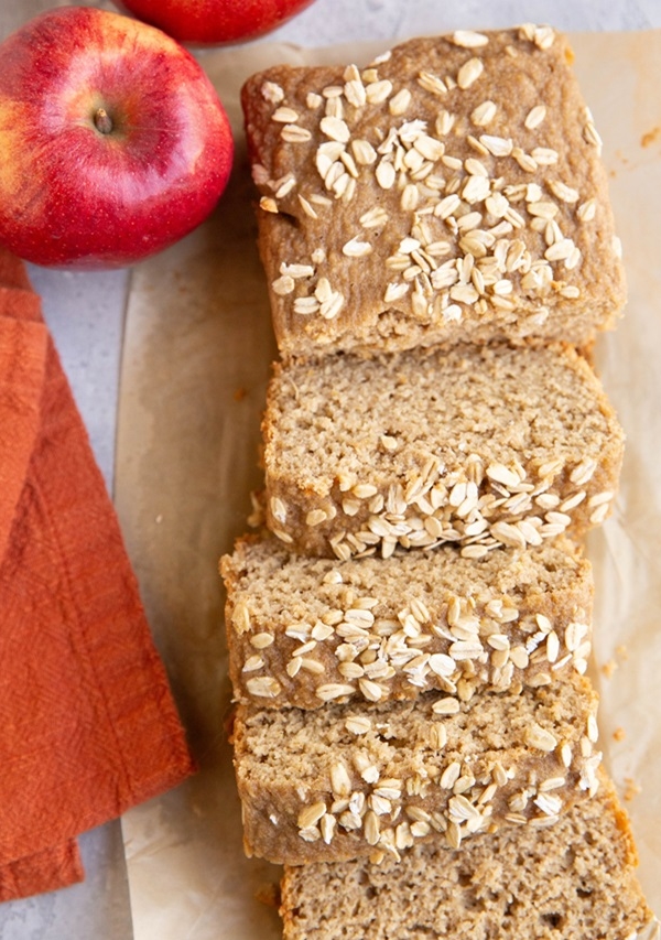 Loaf of applesauce oatmeal bread cut into slices with fresh apples to the side.