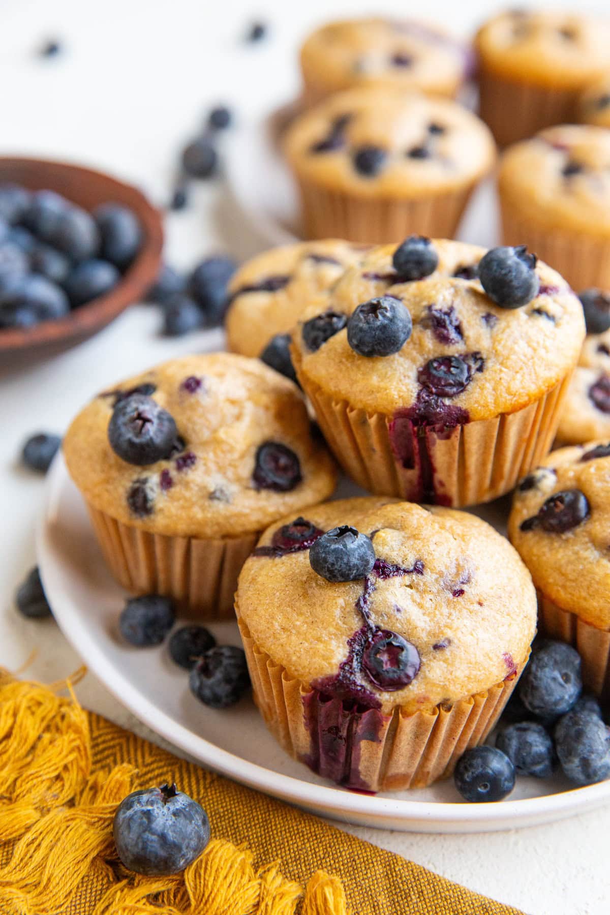 Plate of blueberry muffins with fresh blueberries all around and a golden napkin to the side.