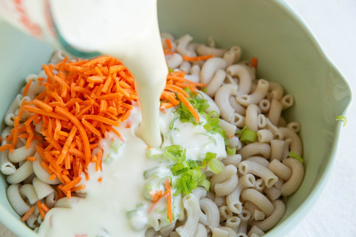 Pouring creamy dressing into a mixing bowl with noodles and veggies to make macaroni salad.