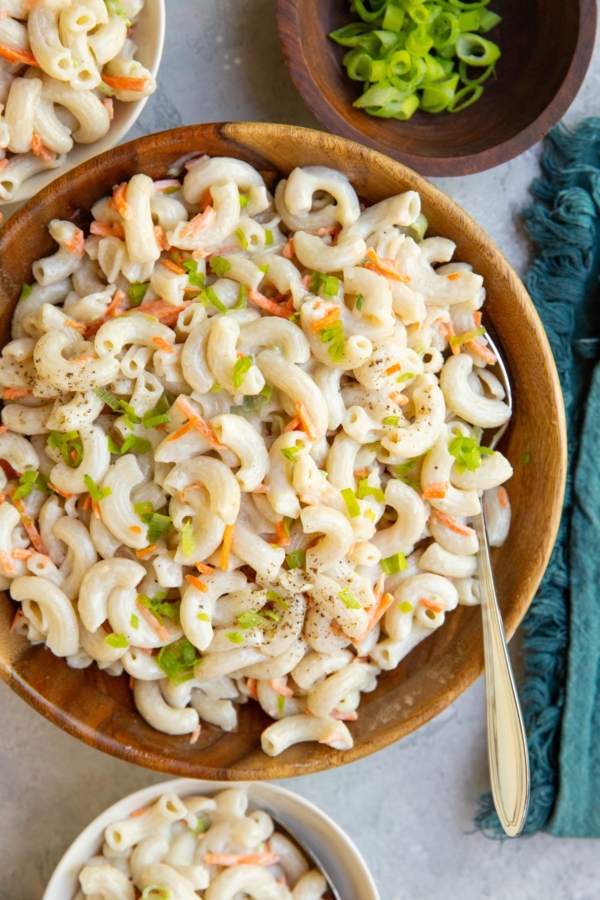 Wooden bowl full of macaroni salad with small bowls of salad and a bowl of chopped green onions