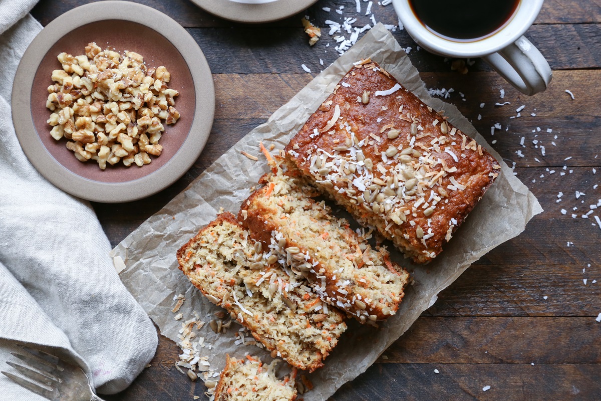 Loaf of morning glory bread cur into slices on a wooden background with a plate of walnuts to the side.