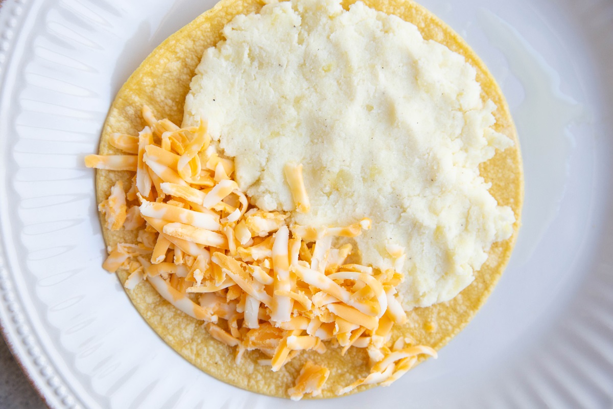 Corn toritlla on a plate with mashed potatoes on one side of the tortilla and grated cheese on the other side.