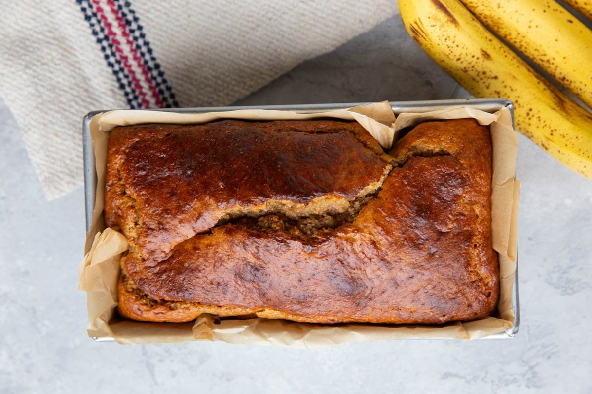 Loaf of banana bread in a loaf pan, fresh out of the oven, with fresh bananas to the side.