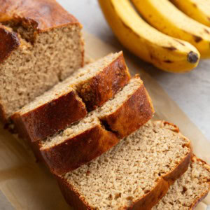 Loaf of high-protein banana bread on a backdrop, cut into slices with bananas in the background.