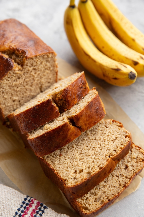 Loaf of high-protein banana bread on a backdrop, cut into slices with bananas in the background.