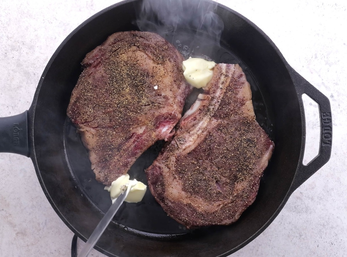 Cast iron skillet with steaks and butter being seared over high heat.