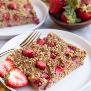 Two slices of strawberry breakfast bars on white plates with the rest of the breakfast bars in the background.