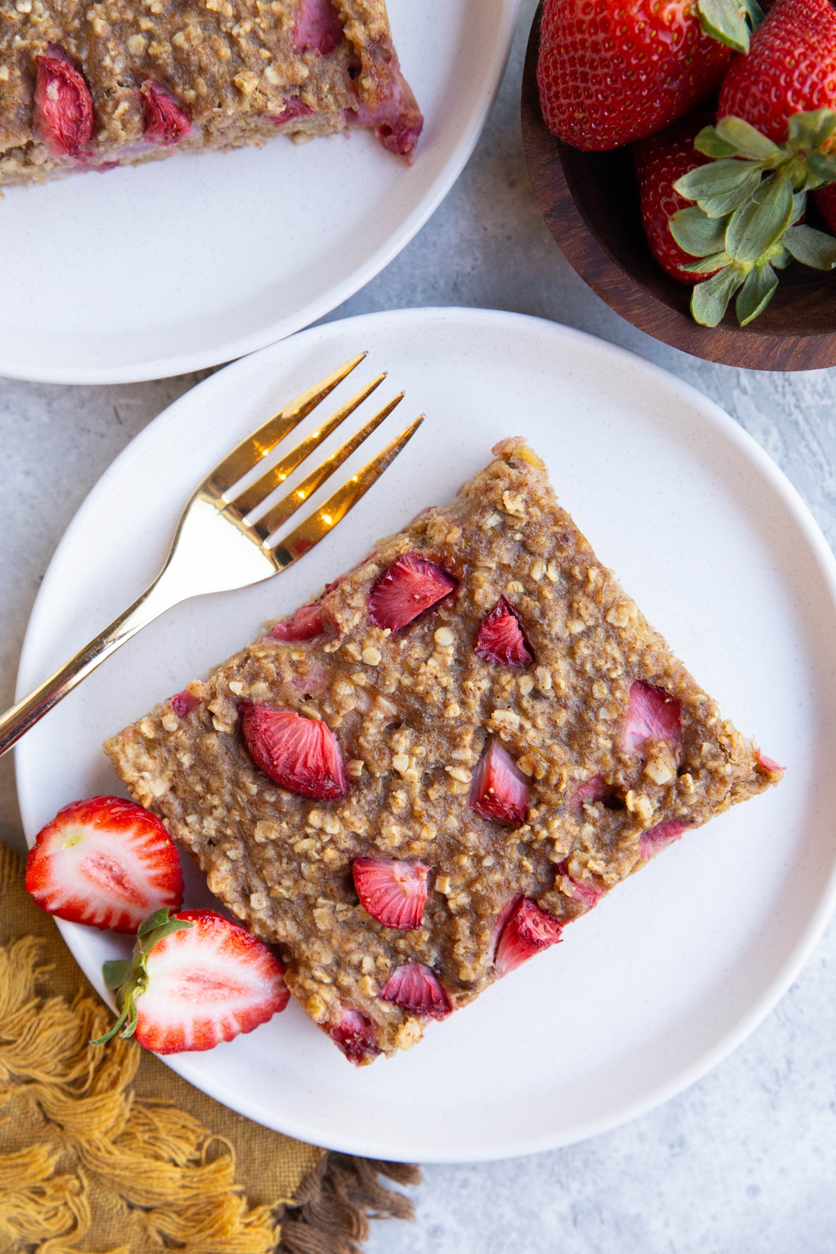 Two white plates with a slice of strawberry breakfast bar and gold forks for eating. A bowl of fresh strawberries and a golden napkin to the side.