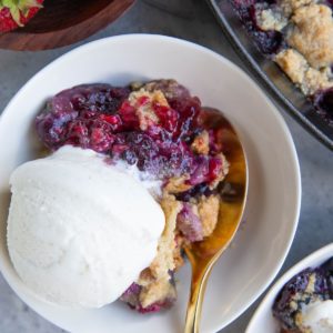 Mixed berry crisp in a white bowl with fresh strawberries to the side.