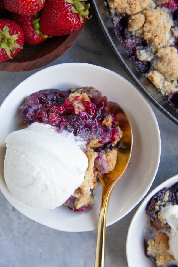 Mixed berry crisp in a white bowl with fresh strawberries to the side.