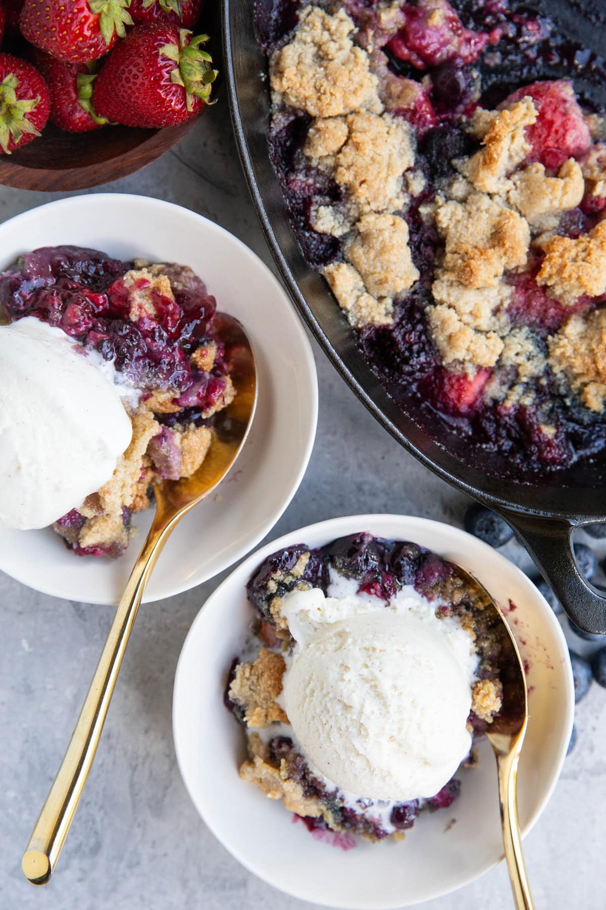 Two bowls of berry crisp with a scoop of vanilla ice cream on top and the rest of the crumble in a baking dish to the side.